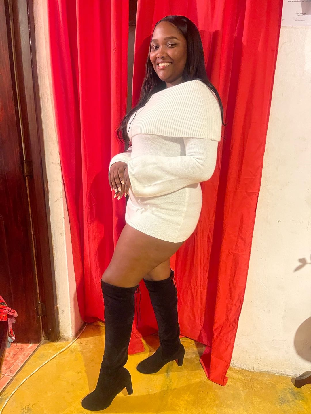 A woman from the Dominican Republic in a white dress and boots, posing in front of a red curtain like a live stream set
